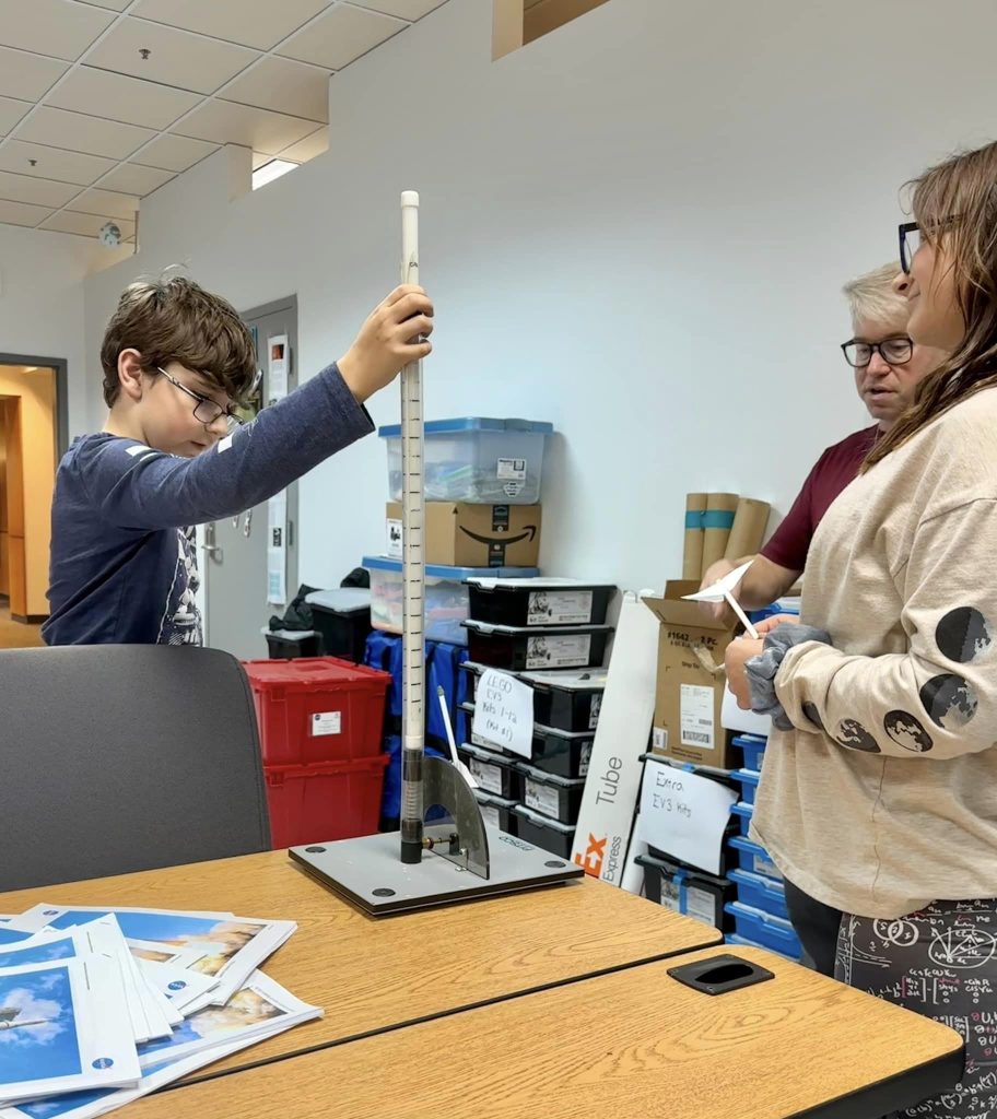 A student raising a tall clear tube on a lab stand while two others look on.